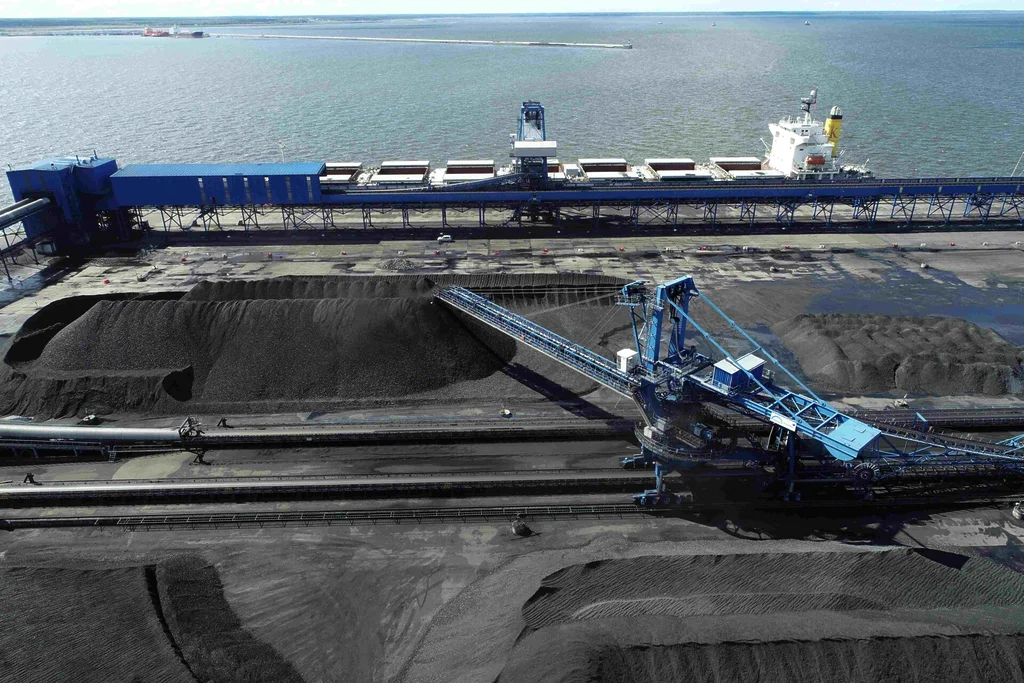 Large coal stockpiles with conveyor and stacking equipment beside a bulk carrier at an export terminal.